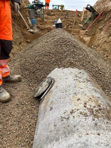 Boden mit einem großen, teilweise freigelegten Betonrohr in einer Baugrube.